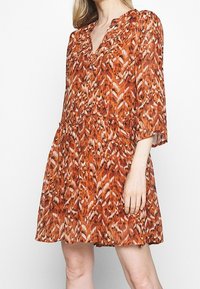 Femme portant une robe courte, ample, avec un imprimé animal abstrait orange et marron et des manches trois-quarts.
