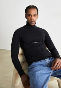Maglione a collo alto nero con polsini e orlo a coste, caratterizzato dal logo bianco "Calvin Klein Jeans" sul davanti. Indossato con jeans in denim azzurro chiaro.