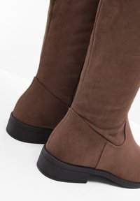 Stivali alti in suede marrone con tacco basso a blocco, dotati di zip laterale e texture liscia. Il design è aderente intorno al polpaccio.