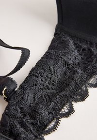 Coque de soutien-gorge en dentelle noire avec motifs floraux et bretelle réglable sur fond clair.