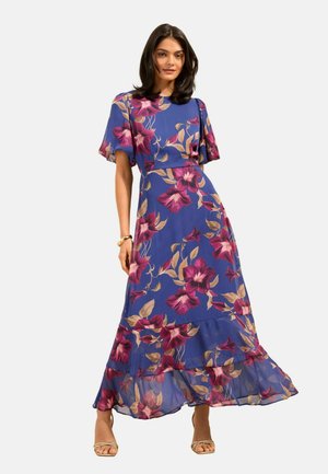 Nő kék, virágos maxiruha viselésével, rövid pufó ujjakkal és bézs pántos sarokcipővel, egyszerű világos háttér előtt pózolva.