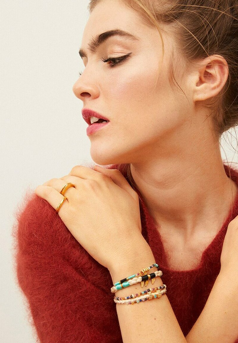 Bague en or au doigt. Bracelets en perles colorées avec des accents turquoise, noirs et multicolores. Le modèle porte un pull rouge en peluche.