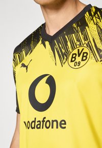 Gul fodboldtrøje med sorte detaljer. Har v-hals, logo på brystet og abstrakte mønstre på skuldrene. Lavet af åndbart stof.