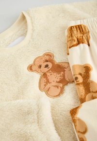 Kremo spalvos fliso džemperis su ruduo teddy lokiuko aplikacija. Atitinkamos kelnės turi panašų teddy lokiuko raštą minkštose, šiltose spalvose.