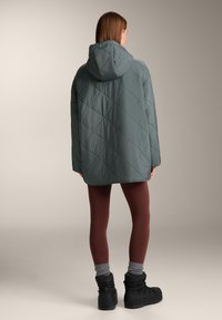 Quilted, hooded jacket in muted teal, paired with brown leggings and black boots. Features a boxy silhouette and textured fabric.