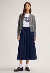 Grijze gebreide cardigan met zilveren knopen, wit t-shirt met een blauw hartmotief en marineblauwe geplooide rok. Zwarte loafers met gouden accenten.