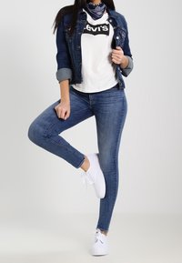 Chaqueta de mezclilla, camiseta blanca con logo negro, jeans ajustados azul oscuro y zapatillas blancas. Los accesorios incluyen un pañuelo azul con estampado.