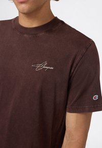 Hombre vistiendo una camiseta Champion de color marrón oscuro con logo bordado blanco en el pecho y pequeño parche de Champion en la manga izquierda.