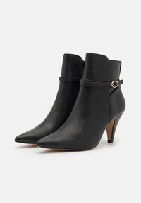 Botines de cuero negro con punta afilada y tacón stiletto. Presentan una correa decorativa con un acento dorado alrededor del tobillo.