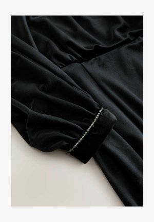 Schwarzes Samtkleid mit langen Ärmeln, das an der Taille geraffte Details und verzierte Manschetten mit einer Reihe von kleinen, glänzenden Akzenten aufweist.