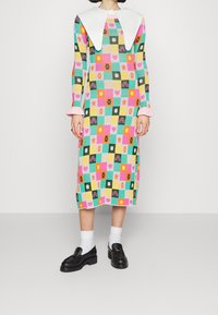 Robe en tricot avec un motif patchwork vibrant dans des couleurs pastel, agrémentée d'un col Peter Pan blanc et de poignets froncés roses. Mocassins noirs en dessous.