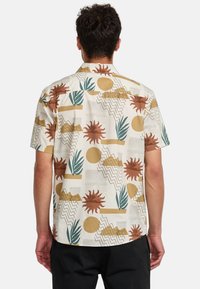 Camicia a maniche corte in tessuto leggero, con una base beige e motivi astratti di sole, montagne e piante in tonalità ruggine e verde.
