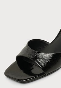 Μαύρο παπούτσι mule από δέρμα με πατέντα, ανοιχτό δάχτυλο, φαρδιά λουρίδα και τετράγωνη μύτη σε απλό ανοιχτό φόντο.