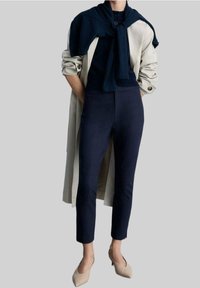 Trench-coat beige clair sur un haut marine, avec un pull marine noué autour des épaules ; un pantalon fitted marine et des escarpins nude complètent la tenue.