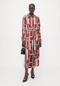 Abito camicia lungo in bordeaux con motivi geometrici bianchi, caratterizzato da maniche lunghe, chiusura con bottoni frontale e una cintura abbinata in vita.