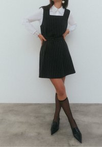 Robe noire à fines rayures avec un col carré et une ceinture assortie ; portée sur une chemise blanche à col. Chaussures noires pointues et bas noirs transparents.