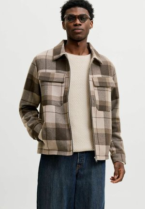 Mann mit runden Brillen, beigem Strickpullover, braun-beiger karierter Jacke mit Brusttaschen und dunkelblauen Jeans, stehend mit einer Hand in der Tasche.