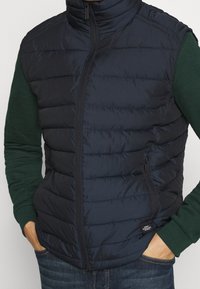 Granatowa pikowana kamizelka z wysokim kołnierzem i zamkiem błyskawicznym, wyposażona w poziome przeszycia, teksturowaną tkaninę oraz boczną kieszeń na przechowywanie.