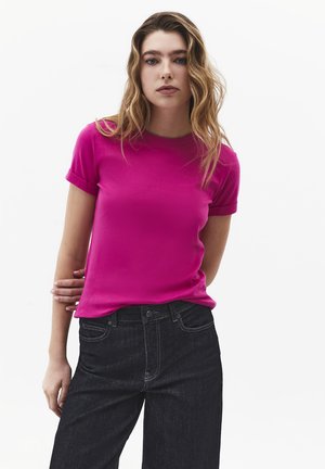 Jonge vrouw met golvend haar, gekleed in een felroze shirt met korte mouwen en donkere spijkerbroek, staand tegen een witte achtergrond.