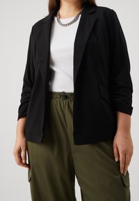 Černé sako s přizpůsobeným střihem, jednoknopovým zapínáním a dvěma bočními kapsami, nošené přes bílý žebrovaný top a zelené kalhoty s elastickým pasem.