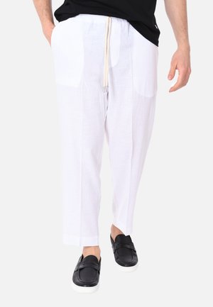 Uomo che indossa pantaloni bianchi con coulisse e scarpe nere senza lacci che cammina davanti a uno sfondo semplice.