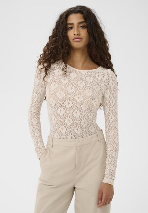 Spitzen-Bodysuit mit langen Ärmeln in sanftem Beige mit floralen Mustern. Kombiniert mit hoch taillierten beigefarbenen Hosen mit seitlichen Taschen und glattem Stoff.