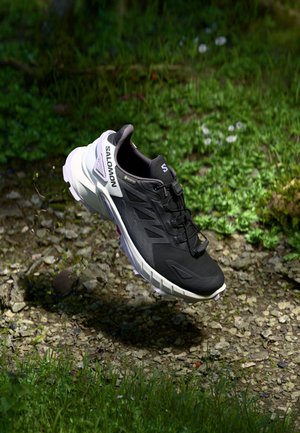 Czarno-biały but do biegania trail Salomon z Gore-Tex, unoszący się nad żwirem i trawą na leśnym podłożu.