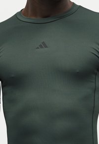 T-shirt de sport ajusté vert foncé avec logo Adidas noir centré sur la poitrine et coutures visibles sur les côtés.