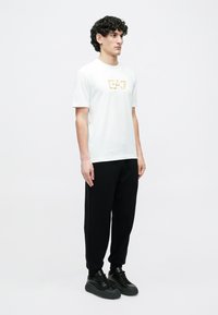 Mladý muž oblečený v bielom tričku s grafickým motívom, čiernych teplákoch a čiernych teniskách stojaci pred jednoduchým bielym pozadím.