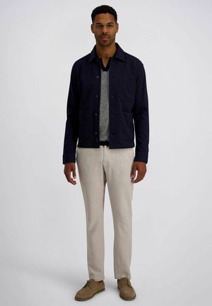 Man staat met ontspannen handen, draagt een donkerblauwe jas, een grijze V-hals trui, beige broek en bruine schoenen tegen een effen achtergrond.