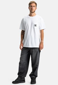 Biała bawełniana koszulka z czarnym logo na kieszeni na piersi, zestawiona z luźnymi, ciemnoniebieskimi dżinsami oraz czarnymi trampkami z teksturowanymi detalami.