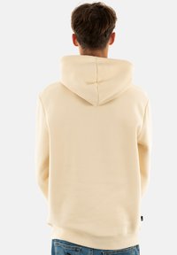 Felpa beige con cappuccio, caratterizzata da una texture liscia e polsini a costine, indossata sopra jeans di denim blu, vista da dietro.