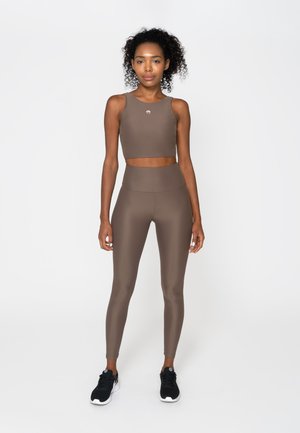 Woman standing wearing brown sleeveless crop top and matching high-waist leggings with black athletic shoes against white background.