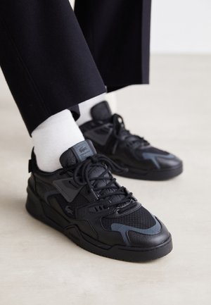 Sneakers Lacoste nere e grigie indossate con calzini bianchi e pantaloni neri su uno sfondo chiaro di pavimento.