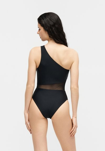 Femme aux cheveux bruns ondulés portant un maillot de bain noir asymétrique avec un panneau en maille transparente à la taille, vue de dos.