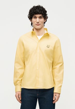 Νέος άνδρας με καστανά σγουρά μαλλιά, που φοράει ένα κίτρινο πουκάμισο με κουμπιά και ένα μικρό ζωάκι λογότυπο στο στήθος και σκούρα τζιν, σε απλό φόντο.