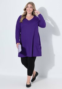 Lange lila Tunika mit V-Ausschnitt, seitlichen Taschen, kombiniert mit schwarzen Leggings und schwarzen Slippers; hält eine helllila Clutch.