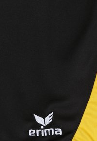 Zwarte sportshorts met een gele zijpaneel, met een geborduurd wit logo "erima" op de linkeronderkant. Gladde textuur van de stof.