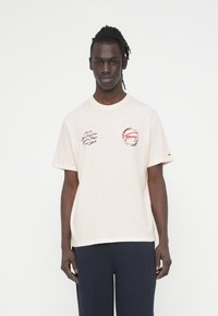 Ανοιχτό μπέζ βαμβακερό T-shirt με αέρινη εφαρμογή, με κόκκινο και μαύρο γραφικό κείμενο και λογότυπο στο στήθος. Στρογγυλή λαιμόκοψη και κοντά μανίκια.