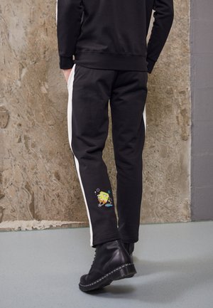 Person trägt schwarze Hosen mit weißen Seitenstreifen und eine kleine SpongeBob Schwammkopf-Grafik nahe dem Unterschenkel, kombiniert mit schwarzen Stiefeln.