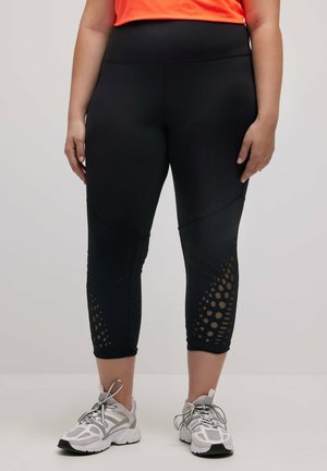 Schwarze Leggings mit hohem Bund, die durch perforierte Akzente am Saum und eine glatte, dehnbare Textur bestechen. Tragen mit Sportschuhen.