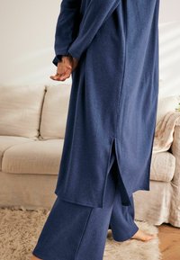 Tamsiai mėlynas laisvalaikio kostiumas, sudarytas iš ilgo švarko su šoninėmis išpjovomis ir laisvai krentančių kelnių, pagamintas iš minkštos medžiagos su jaukia tekstūra.