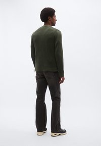 Zelen pulover z rebrasto tkanino, ravne črne kavbojke in temni supergi. Strukturna zasnova, teksturirana tkanina in preprosti poudarki. Pogled od zadaj.