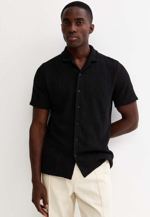 Uomo che indossa una camicia nera a maniche corte con bottoni e pantaloni chiari, in piedi contro un muro bianco con una mano dietro la schiena.