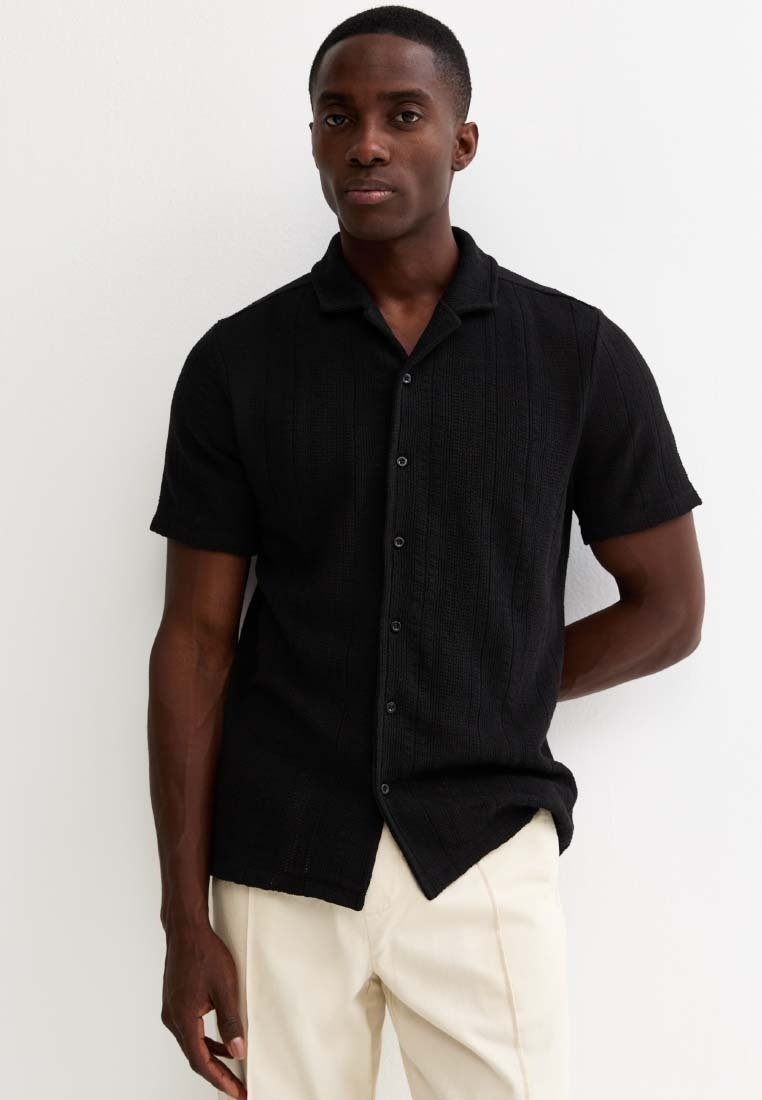 Man draagt een zwart kortärmelig overhemd met knopen en lichtgekleurde broek, staand tegen een witte muur met één hand achter zijn rug.