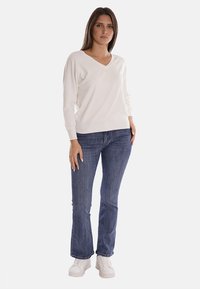 Maglione bianco a V con maniche lunghe, abbinato a jeans in denim blu a gamba larga e sneakers bianche, con un fit casual e rilassato.