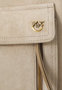 Beige σουέντ τσάντα με καπάκι που διαθέτει λεπτομερή ραφή, χρυσό κούμπωμα σε σχήμα πουλιού και μερικώς ορατό χρυσό φερμουάρ με δερμάτινο χερούλι.