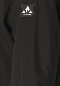 Zwarte jassenstof met een gemêleerde afwerking, voorzien van een vierkante patch met een wit berglogo en het woord "WHISTLER" eronder.