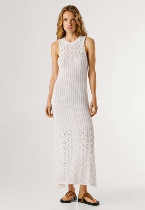 Vrouw met lang, krullend haar, gekleed in een wit, mouwloos gebreid maxi-jurk en bruine sandalen, staand tegen een effen lichte achtergrond.