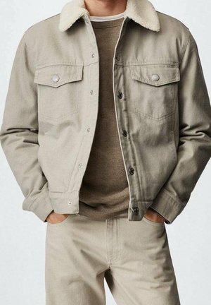 Mand iført beige jakke med sherpa-krave over brun sweater og beige bukser med hænderne i lommerne, vist fra nakke til midt på låret.